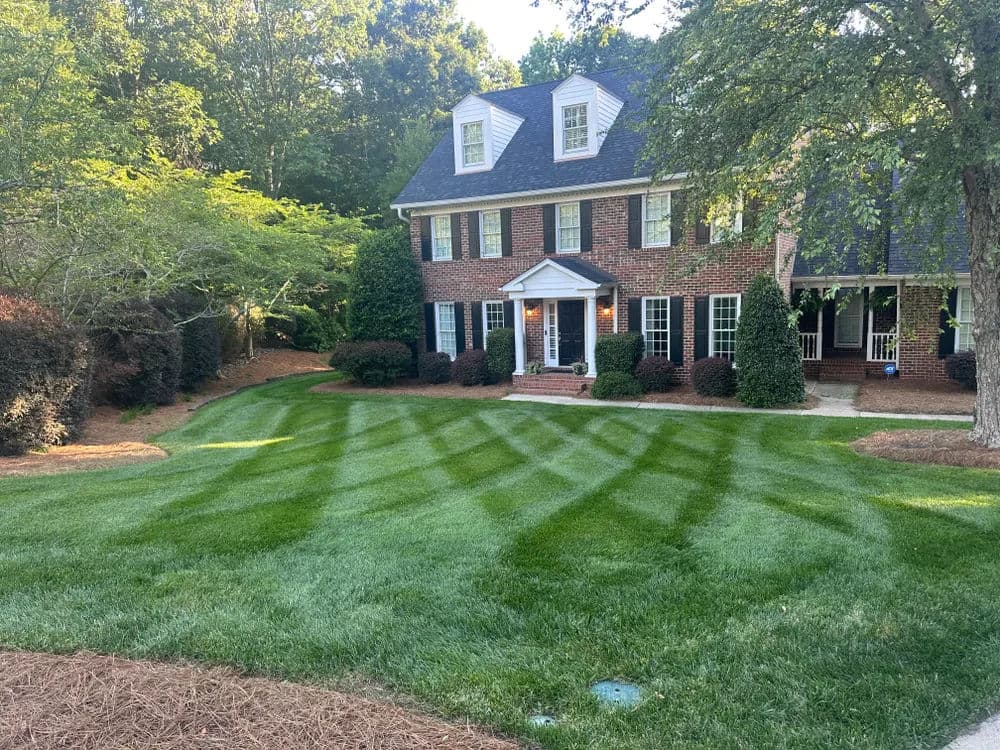 Lush green lawn with striped pattern in front of a brick house surrounded by trees.