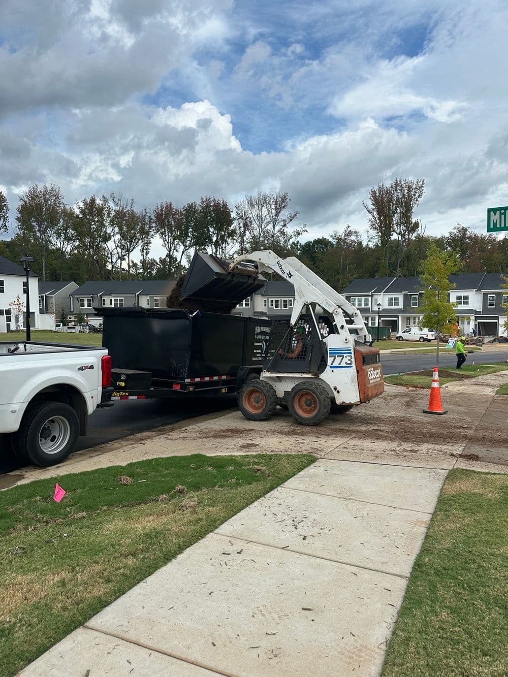 Skid steer loader dumping debris into a large dumpster on a residential street.