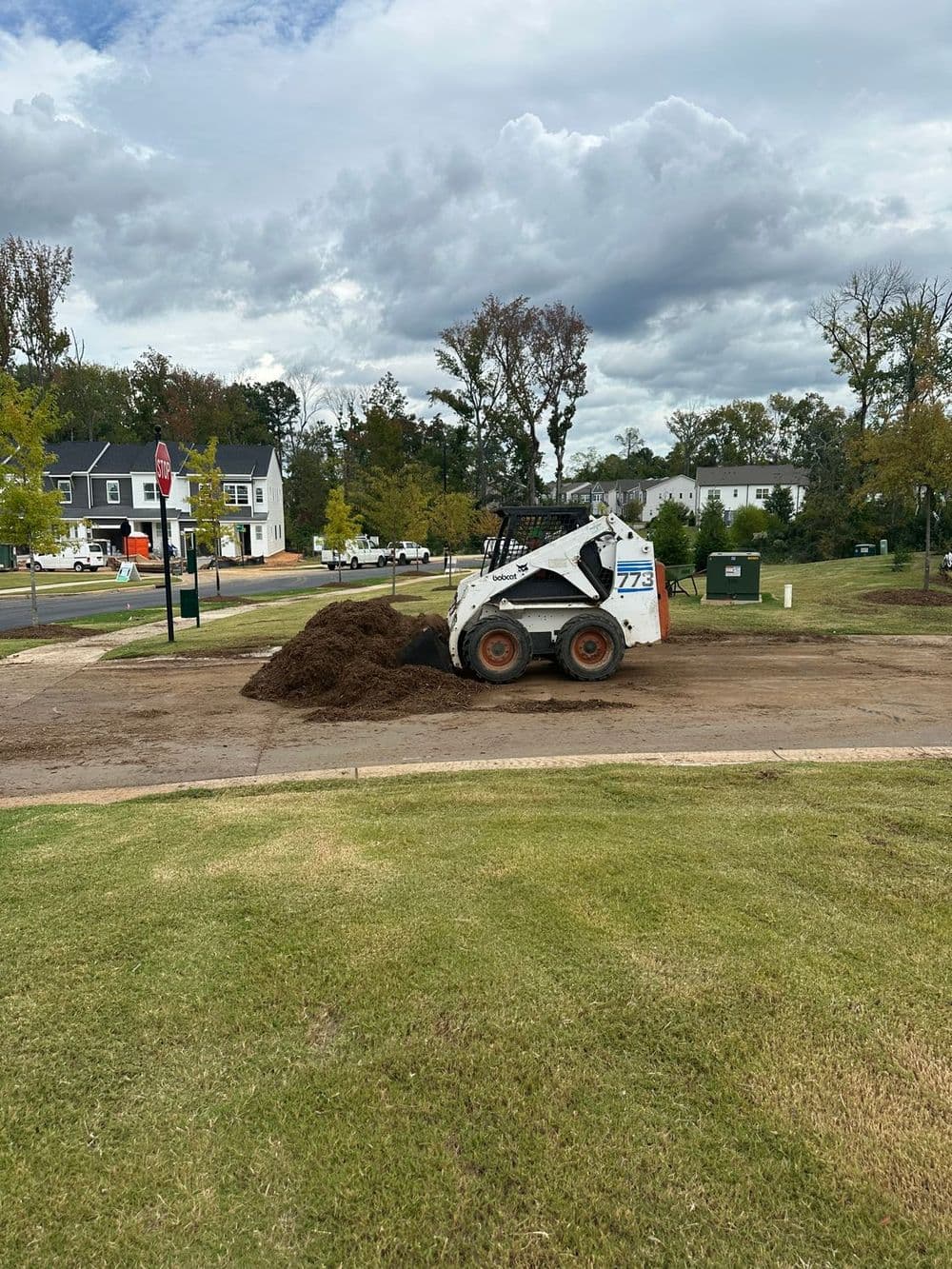 Construction site with a skid steer loader moving mulch under a cloudy sky.