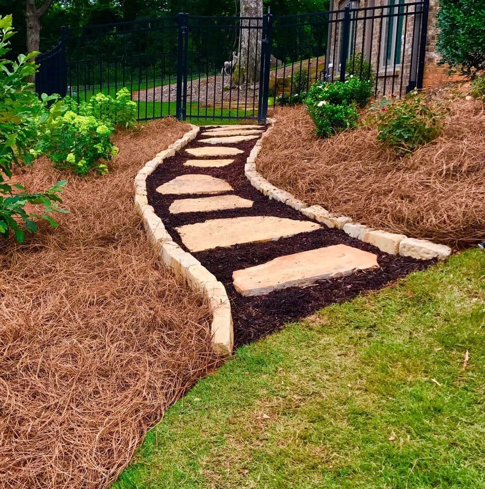 Curved stone pathway bordered by mulch and greenery leading to a gated area.
