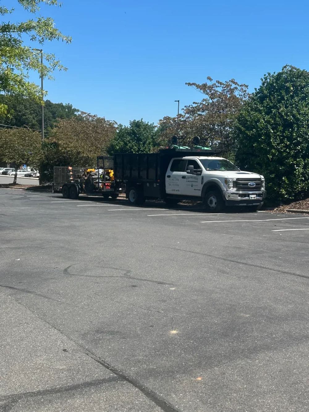 Truck with trailer parked in a sunny lot surrounded by trees and greenery.