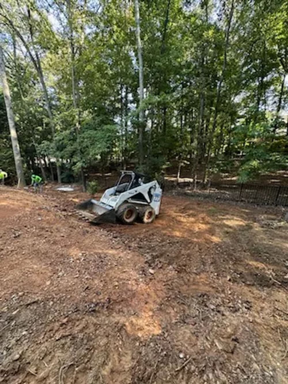 Bobcat skid steer working on landscaping in a wooded area with workers in the background.