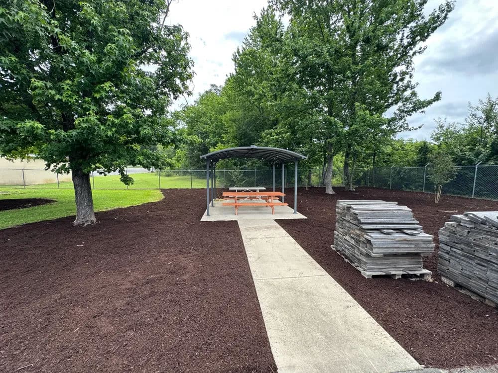 Park area with a picnic shelter, wooden benches, and mulch landscaping surrounded by trees.