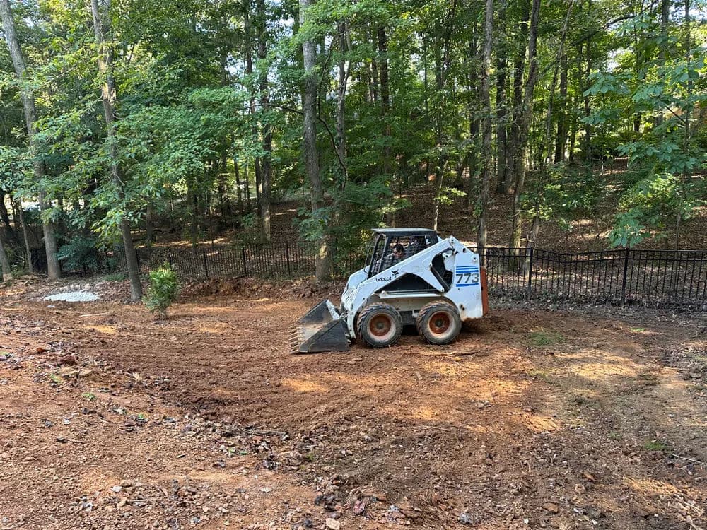 Bobcat skid steer clearing land in wooded area with black fence in the background.