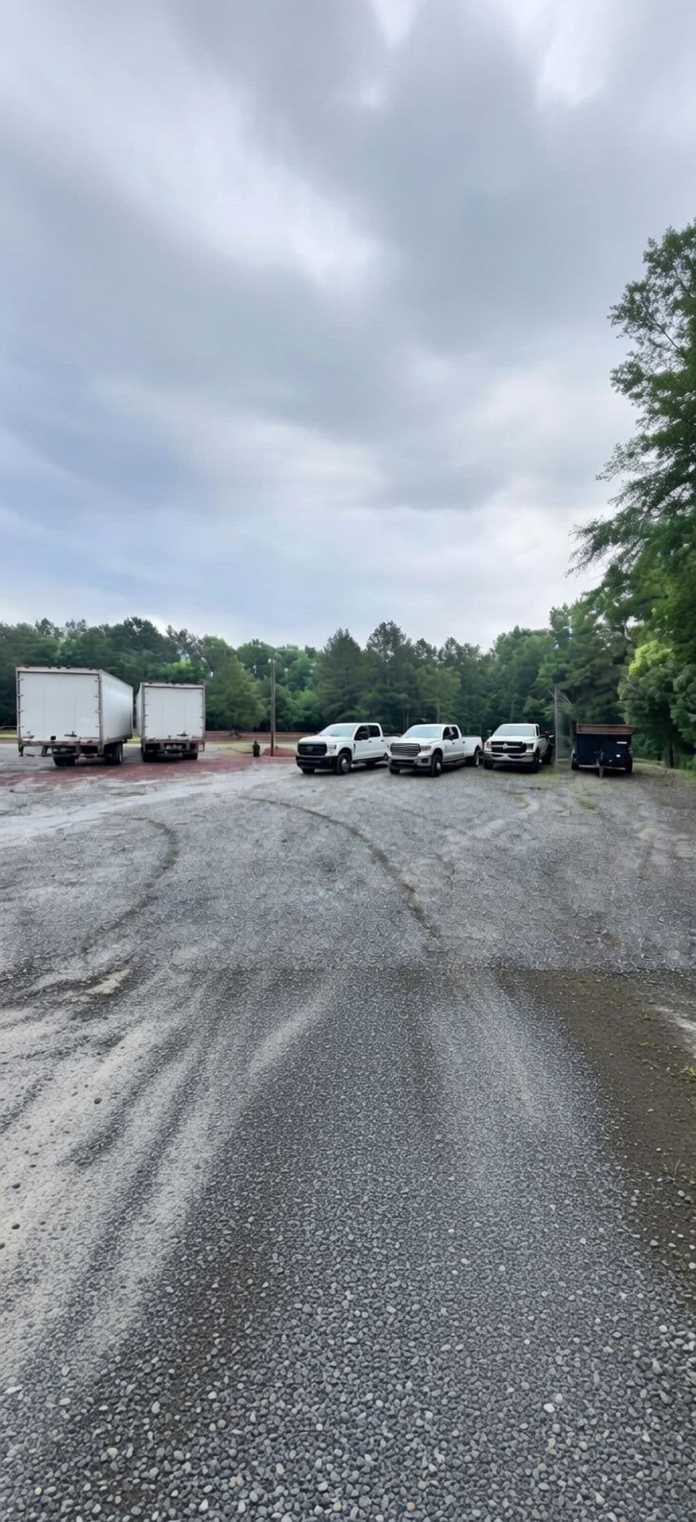 White trucks parked in a gravel lot under cloudy skies surrounded by trees.